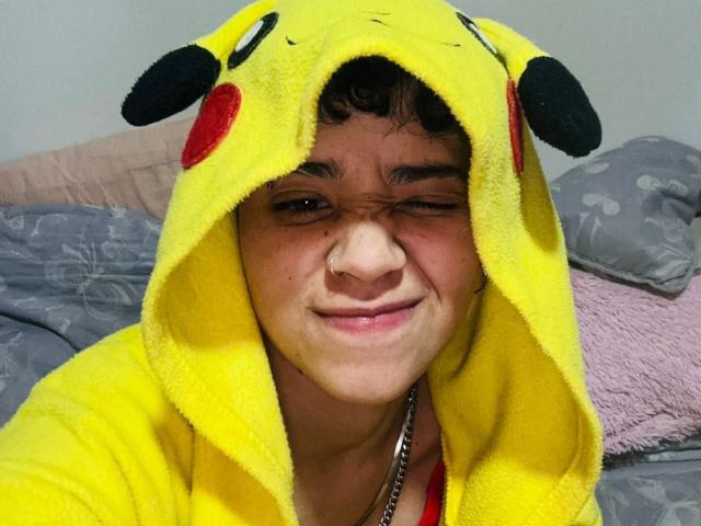 JadeRayden6 has a mischievous smile with one eye closed, wearing a yellow Pikachu hoodie with black ears and red circles. His curly dark hair is visible under the hood. He has a nose ring and a silver chain around his neck. The hoodie's texture is soft and plush. The background includes gray pillows and a pink pillow. The image focuses on JadeRayden6's playful expression and Pikachu costume. Slightly blurry.