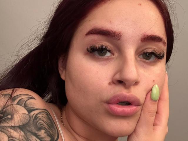 LucieRed has dark red hair, green nails, and a tattoo on her shoulder. Her lips are slightly parted, showing her pinkish lips. She's touching her cheek with her green-nailed hand.