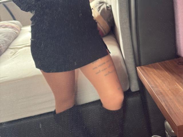 Dirtyheike-hot's bare thighs in a black mini-dress, showing a tattoo on her right thigh. She's wearing black knee-high socks, standing next to a bed and wooden table.