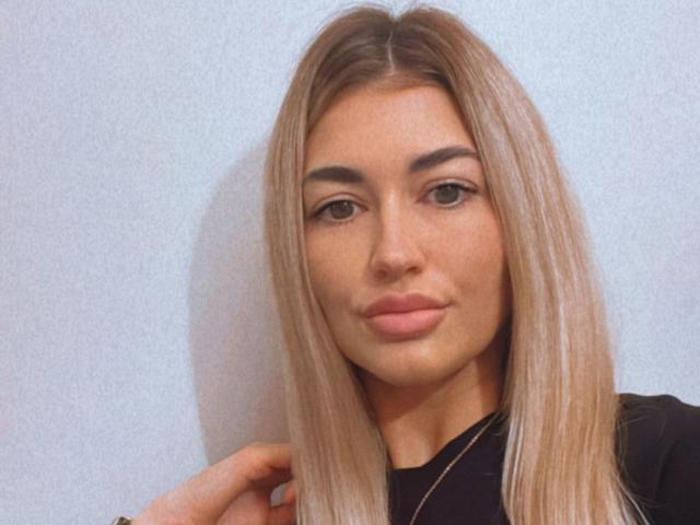 SoniaaBlond tem cabelo loiro liso, sobrancelhas grossas e lábios rosados e carnudos. Ela está vestindo uma blusa preta com um colar fino. Seus olhos estão ligeiramente semicerrados..