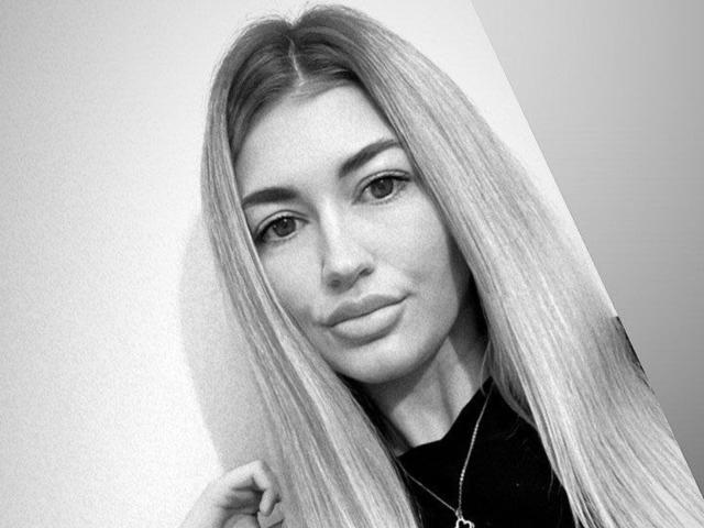 SoniaaBlond with long, straight blonde hair, light skin, and full lips, wearing a black shirt, looking at the camera with a slight smile, grayscale, selfie style.