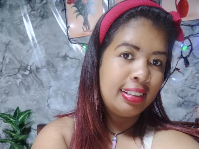 MariahSquirting69 tem pele morena, batom vermelho e cabelo ruivo com uma faixa de cabelo vermelha, vestindo uma blusa branca com um colar roxo, sorrindo com os dentes à mostra..