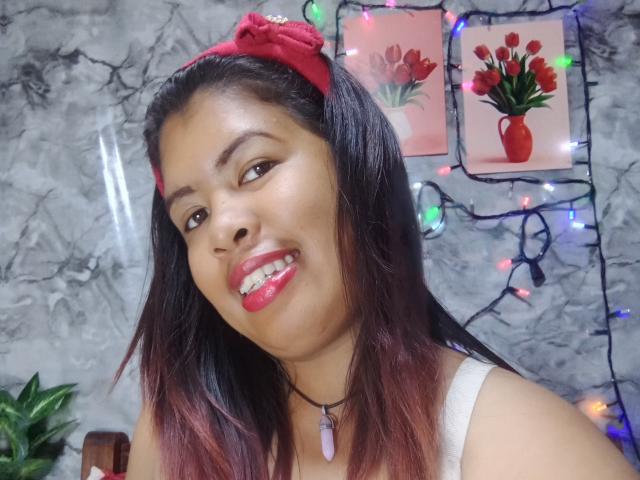 MariahSquirting69 with dark brown skin, red lipstick, and red headband, smiling, wearing a white top, and a pink crystal necklace, in a selfie with red-tinted hair.