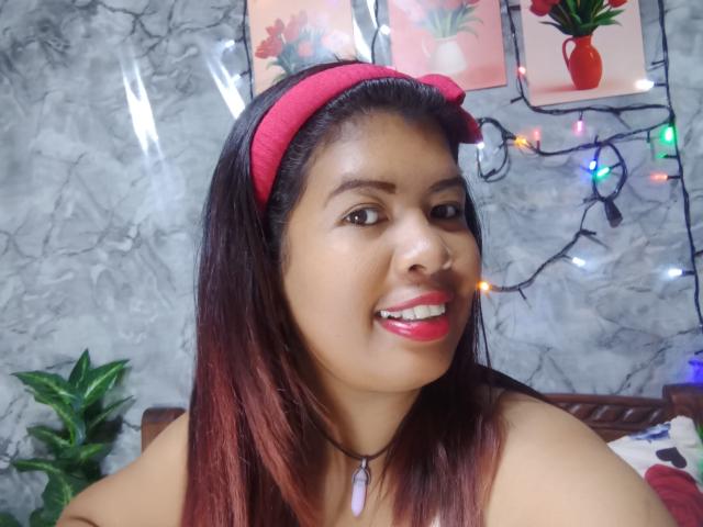 MariahSquirting69 with brown skin, red lipstick, and straight dark red hair, wearing a red headband, smiling, and a purple necklace, in a room with colorful lights.