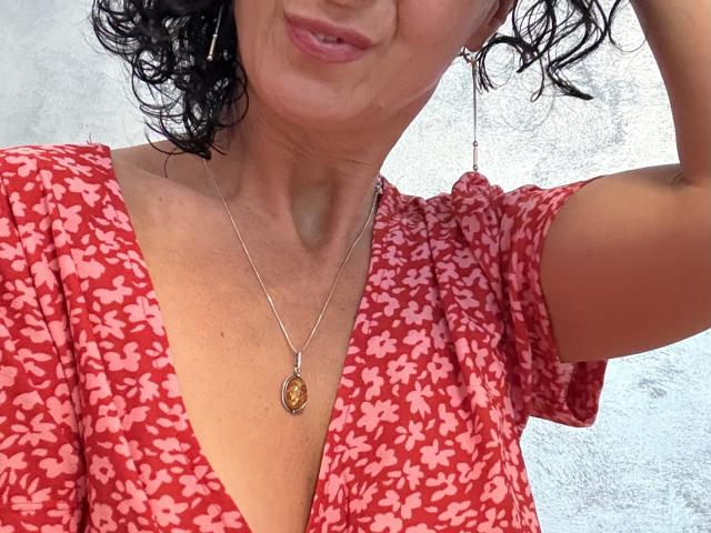 NikHotty with curly black hair, wearing a red floral dress, showing her chest and a silver necklace with a pendant, her nipples slightly visible, smiling subtly.