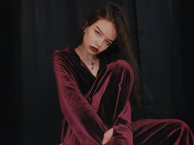 SweetyDeviliya with long dark hair, wearing a red velvet dress, sitting with arms and legs crossed, deep brown eyes, dark red lipstick, and a silver necklace.