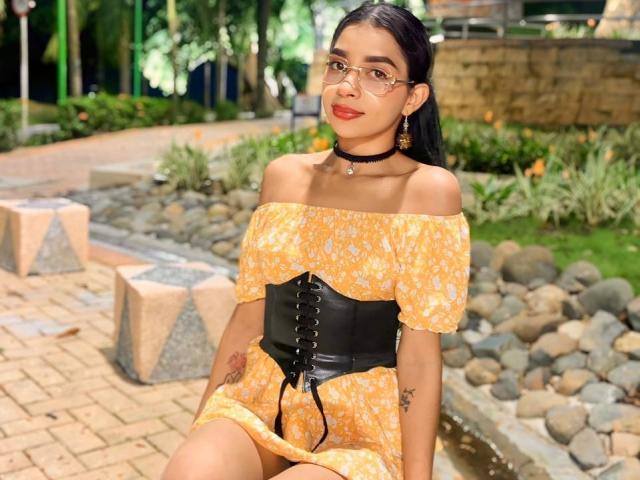RosieRousse, com seios pequenos, usando óculos, um vestido amarelo com um ombro descoberto, um corpete preto, um colar preto e brincos, sentada ao ar livre, sorrindo, com cabelos longos e pretos..