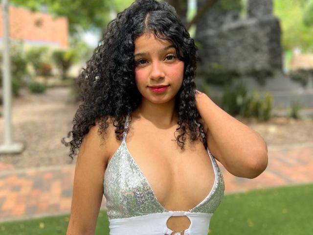 BellainLove is a young woman with medium brown skin and curly black hair, standing outdoors. She wears a silver sequin bikini top with a cutout below her breasts, revealing her cleavage. Her right hand touches her hair, and she has red lipstick on. The green grass and blurred park-like background are out of focus. Her expression is calm, and she has a slight smile. The image has bright, natural lighting.