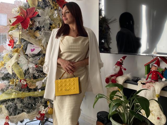 Gala Malkova, con cabello castaño, vestido blanco y chaqueta blanca, sosteniendo un bolso acolchado amarillo, está de pie junto a un árbol de Navidad decorado con adornos rojos y dorados, y pequeñas figuras navideñas..