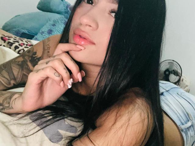 Mady69X has long black hair, pink lips, and tattooed arms, lying on a bed with her finger on her chin, showing off her painted nails and cute smile.