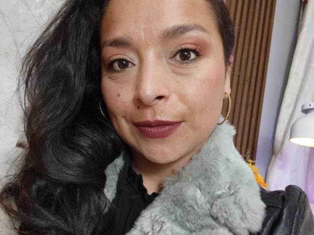 StangelarRivera has medium brown skin, long black hair, small dark moles on her face, wearing purple lipstick, gold hoop earrings, and a gray fur coat, smiling softly.