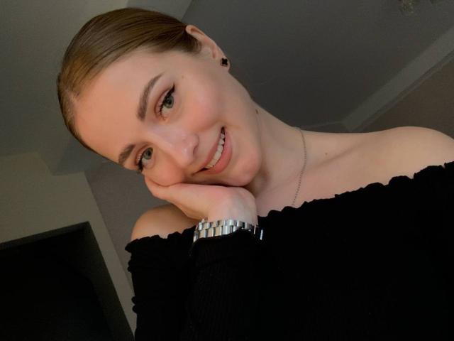 MoonBloom with brown hair in a ponytail, smiling, resting her chin on her hand, wearing a black off-shoulder top, silver watch, and small black earrings.