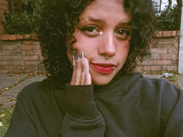 LunaKeller with curly black hair, nose ring, and red lipstick, wearing a black hoodie, touching her cheek with long, silver-painted nails, looking at the camera.
