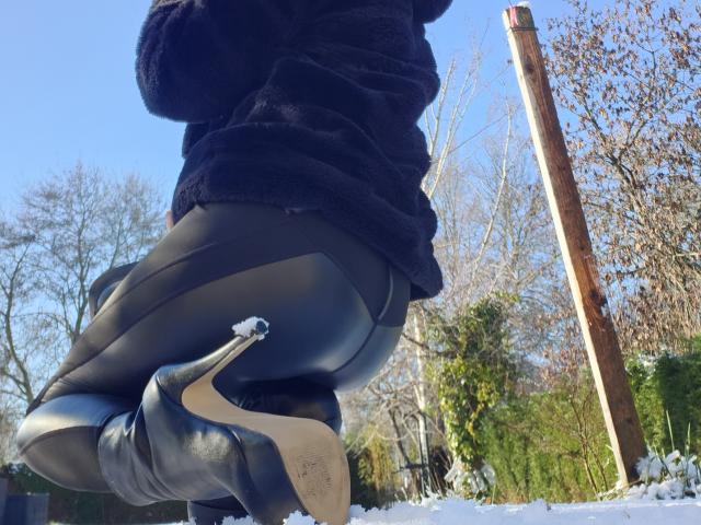 LilouLove squats in a snowy outdoor setting. She wears black leather pants and black stiletto heels with visible wear on the soles. A small snowball is on the heel. LilouLove's dark, fuzzy jacket is partially visible. The background includes leafless trees and a wooden pole. The image has clear, bright lighting. The composition focuses on LilouLove's lower body and the texture of her clothing and shoes. The overall style is candid and bold.