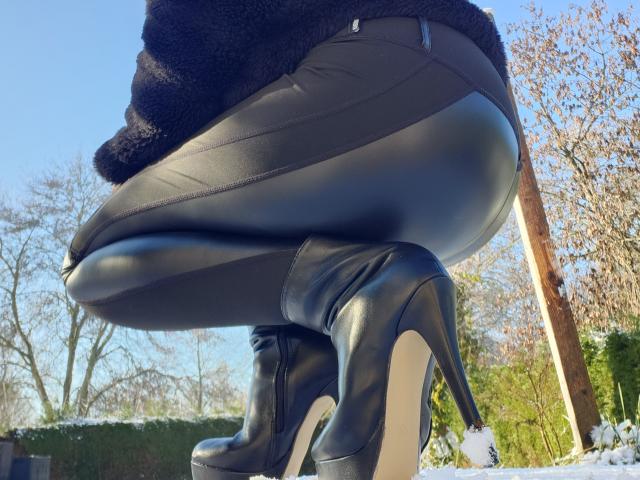 LilouLove is squatting outdoors, wearing black, shiny leather pants that cling to her buttocks and black, high-heeled, platform boots. The boots have a glossy finish and are slightly elevated. She is also wearing a black, fuzzy jacket. The image is taken from a low angle, focusing on her lower body. The background includes leafless trees and greenery, with sunlight casting shadows. The image uses bright, natural light. The overall composition emphasizes the leather texture and the height of the boots.