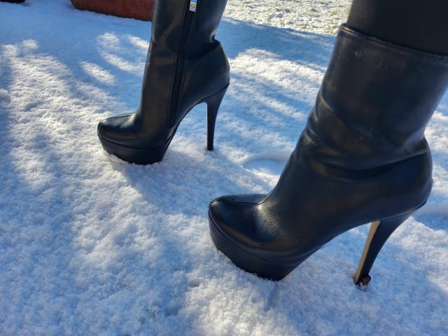 Black leather high-heeled boots with platform soles and stiletto heels stand on a snow-covered ground. The boots are slightly shiny, reflecting light. Snow is textured with small, uneven clumps. The boots' zippers are visible, and the heels are gold-colored. The light source is from the top right, casting shadows on the snow. The boots dominate the composition, with a focus on their glossy texture and platform height. LilouLove's legs are partially visible, indicating she is wearing the boots.