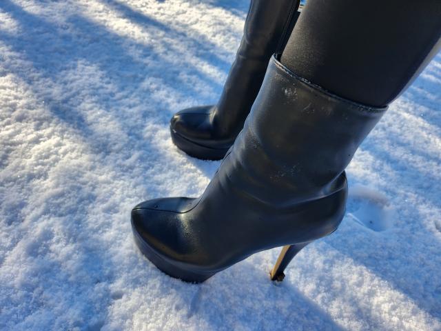 Black leather boots with a high heel and platform sole stand in a snowy landscape. The boots are slightly worn, showing light scuffs on the surface. The snow is textured with small, uneven mounds. Light shadows from the boots fall diagonally across the snow. The boots are positioned in the lower half of the image, slightly apart, with the heel of the right boot slightly higher than the left.