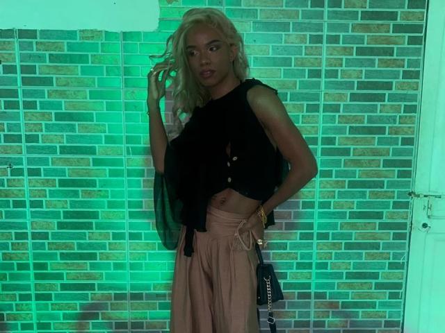 CandyTemptationI, con cabello rubio, top negro sin mangas, abdomen expuesto y pantalones rosas, sosteniendo un bolso negro, de pie frente a una pared de ladrillos iluminada con luz verde..