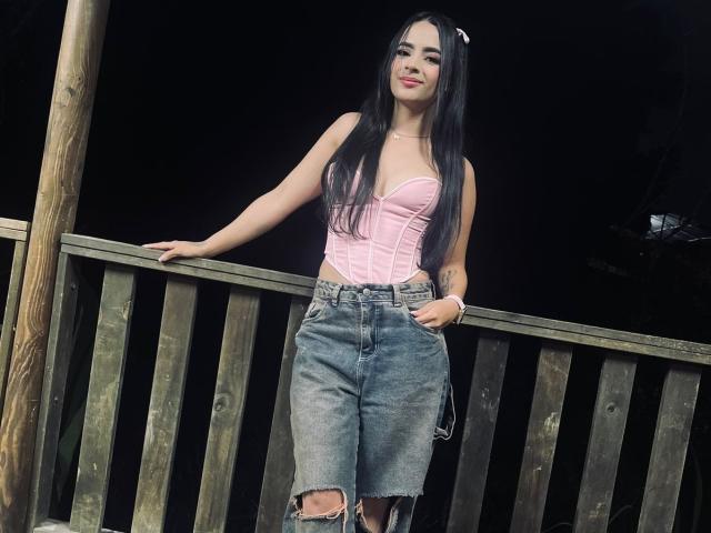 KonanStar with long black hair, pink strapless top, and high-waisted, ripped denim skirt, standing against a wooden railing, smiling with red lipstick.