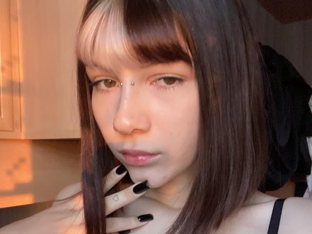 LilyLottie has straight brown hair with white highlights, a nose piercing, black nails, and is touching her chin with a hand near her pouty, glossy lips.