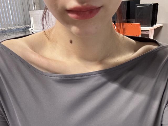 LilyLottie's upper chest, showing a small mole on her neck, with her lips slightly parted, wearing a grey shirt, visible collarbones, and red-painted lips.