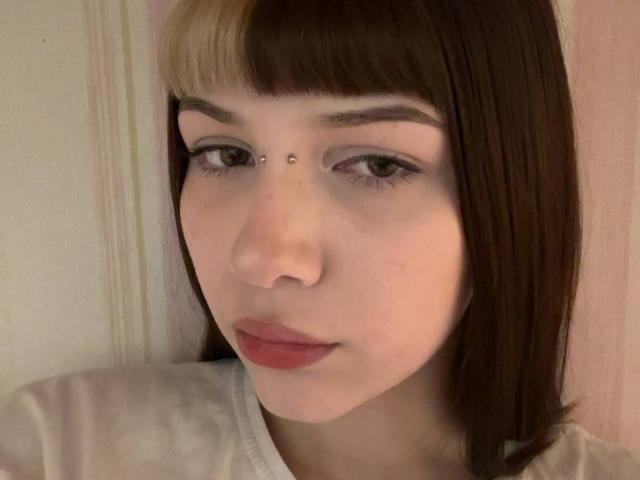 LilyLottie has a pimpled face with a small silver stud on her nose, brown and blonde half-ponytail, and a white shirt, with a neutral expression.