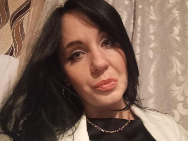 AryCoren has black hair, brown eyes, and pink lipstick. She's wearing a white jacket over a black top with a gold chain. Her ass is not visible.