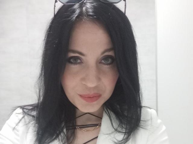 AryCoren with black hair, eyeglasses on head, wearing white shirt, has light skin, gray eyes, and pink lips, looking directly at camera. Boobs partially visible under shirt.