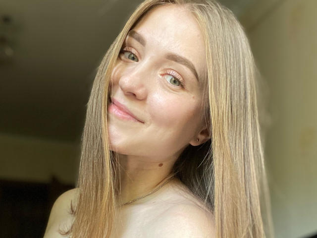 MoonBloom tem cabelo loiro liso, olhos verdes e uma pequena marca de beleza na bochecha direita. Ela está sorrindo suavemente, com um leve inclinação, e está usando um colar de ouro..