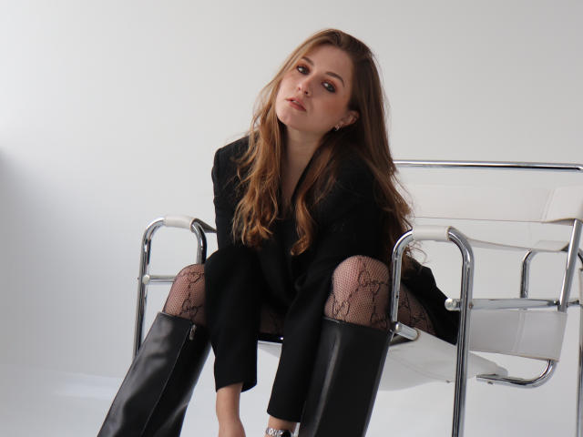 MoonBloom mit langen, braunen Haaren, die einen schwarzen Blazer, Strumpfhosen mit Spitze und schwarze Kniehohe Stiefel trägt, sitzt auf einem weißen Metallstuhl und blickt leicht nach oben..