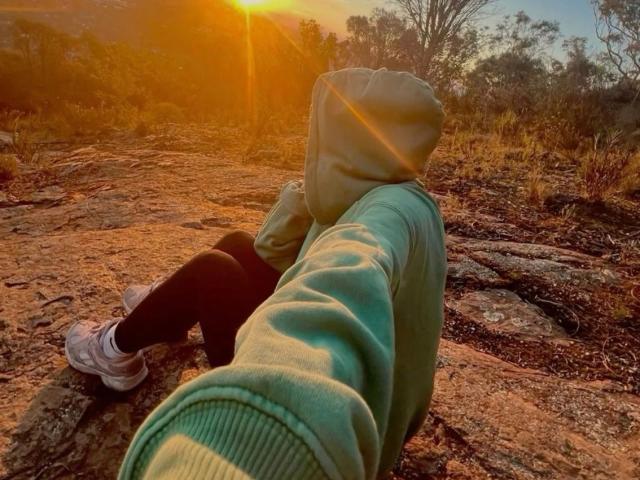 De groene hoodie en zwarte leggings van AmazingYzza, met de billen omhoog, zittend op rotsachtig terrein met uitgestrekte benen, de zon zakt onder in de achtergrond, groene mouwen en witte sneakers, een warme oranje gloed..