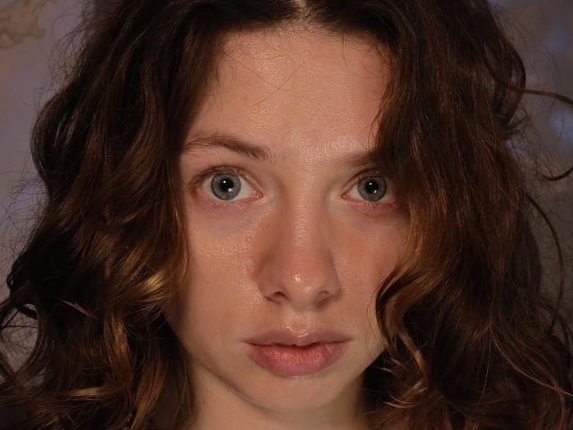 MelodySinclair has wavy brown hair, pale skin, and blue eyes with a slightly sweaty face, looking directly at the camera with a neutral expression.