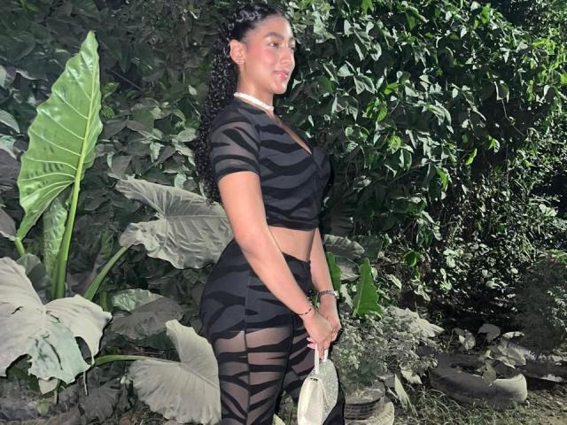 NatalyBlonder with dark curly hair, wearing a sheer black zebra-patterned outfit, showing her curvy ass and big tits, holding a white clutch, standing in a lush green garden.