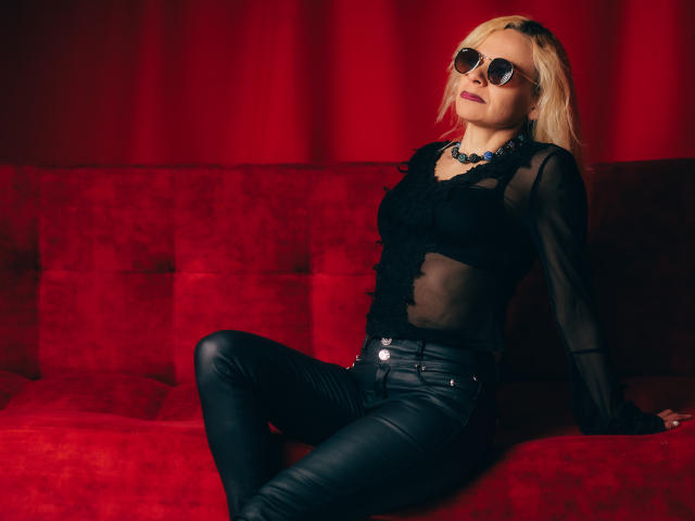 SamanthaJoneX with blonde hair, black sheer top, leather pants, and dark sunglasses, sitting on a red velvet couch, looking confident. Her tits are small, she's sexy.