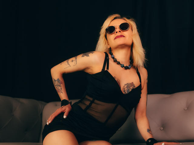 SamanthaJoneX in a black sheer dress, sitting with her ass up, tattoos on arms and chest, blue bead necklace, round sunglasses, blonde hair, dark background, confident pose.