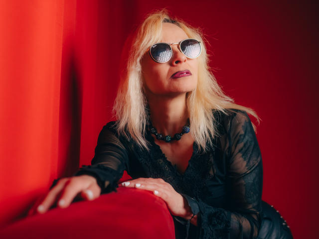 SamanthaJoneX with blonde hair, wearing black lace dress, reflective sunglasses, black necklace, and red lipstick, sitting on red velvet couch, looking up with a confident, sexy expression.