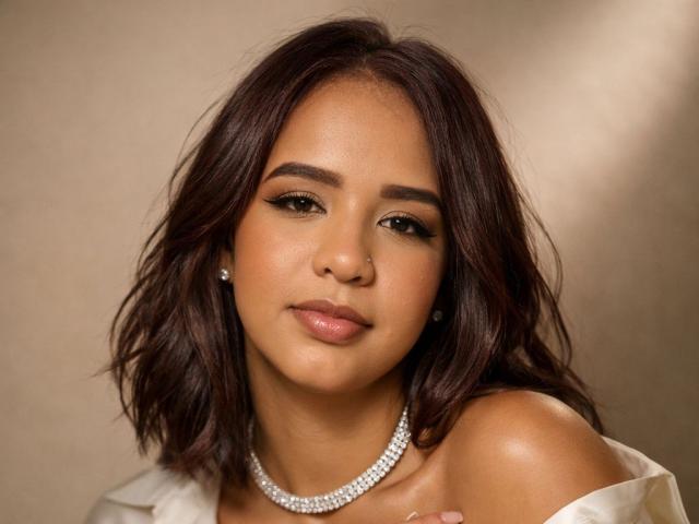 ZoeBlanc with dark brown wavy hair, light brown skin, subtle makeup, and a diamond necklace, looking at the camera with a slight smile, showing her bare shoulders.