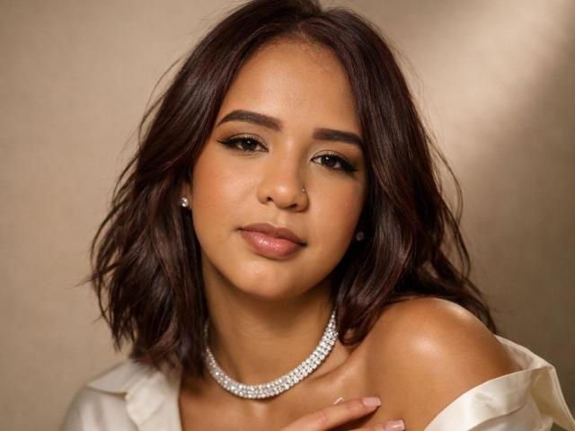 ZoeBlanc has medium brown wavy hair, brown eyes, and tan skin. She's wearing a diamond necklace and a white off-shoulder top, with her tits slightly visible.
