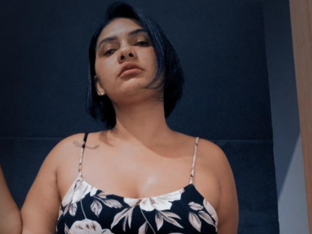 Brun69 with black bob, wearing a floral black and white dress, showing small tits, tattoo on left shoulder, looking down with a slightly pouty expression.
