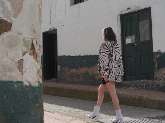 Eliza Canterville camina con una sudadera de estampado de cebra, botas blancas y tanga negra, mostrando su trasero desnudo, por una calle soleada y desgastada por el tiempo..