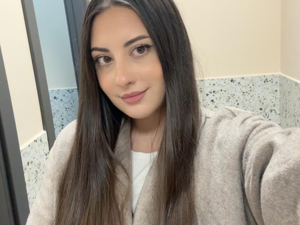 Elyonora has long brown hair, brown eyes, and a subtle smile. She's wearing a gray robe over a white shirt. Her tits are small, and she looks cute.