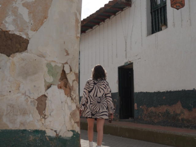 Eliza Canterville, avec ses cheveux bruns bouclés, porte une robe à motifs zébra, et se tient dos à la caméra, montrant ses fesses nues, dans une ruelle rustique et ensoleillée..