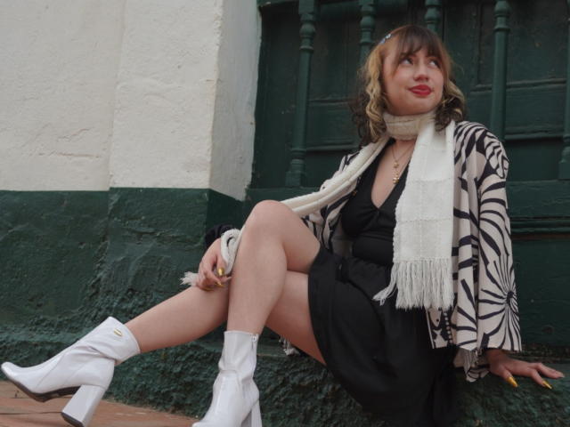 ElizaCanterville sits with legs crossed, wearing a black dress, zebra-patterned jacket, white scarf, and white boots. She has red lipstick, yellow nails, and a relaxed expression.