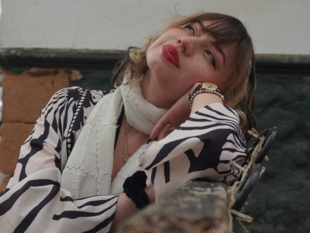 Eliza Canterville, com lábios vermelhos e curvados, blusa com estampa de zebra, lenço branco e pulseiras, apoiada em uma barra de metal enferrujada, olhando para cima com uma expressão ligeiramente entediada..