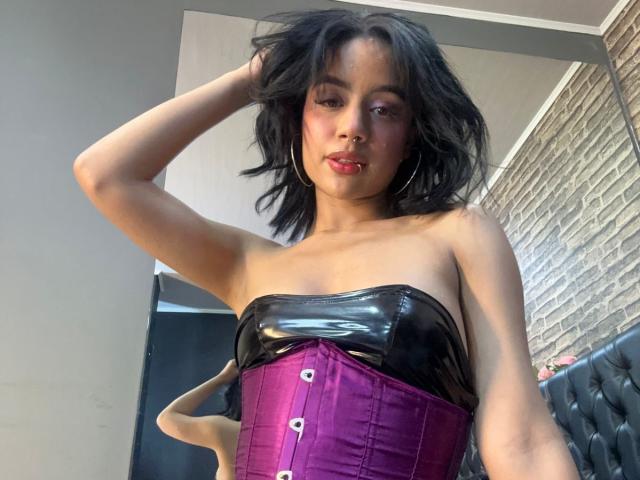 WendyJames with black shiny strapless top, purple corset, and hoop earrings, has short black hair, red lipstick, and is touching her hair with one hand.