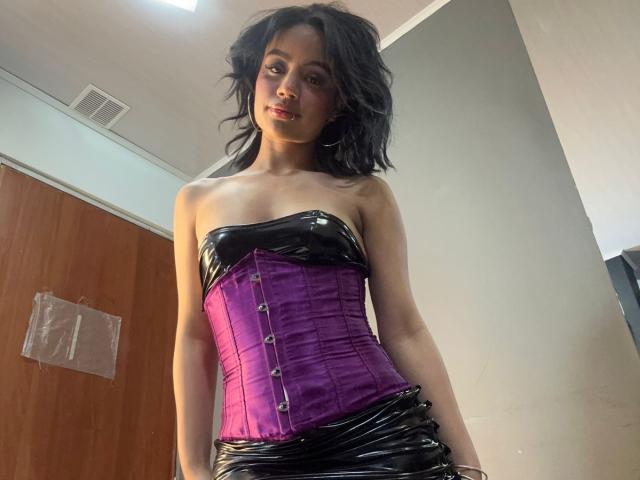 WendyJames with black latex strapless top, purple satin corset, and black latex skirt, standing in room, looking down with a slight smile, wavy black hair.