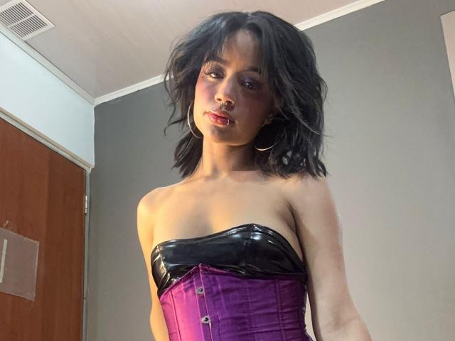 WendyJames with black wavy hair, wearing a purple and black latex strapless corset, showing her tits, and wearing large gold hoop earrings, looking seductively at the camera.
