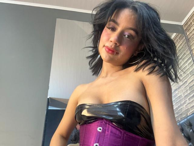 WendyJames with black shoulder-length hair, wearing a shiny black strapless top and purple corset, showing small tits, and a septum ring, smiling slightly.