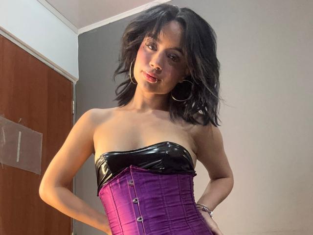 WendyJames with medium tits, wearing a shiny black strapless top and purple corset, black wavy hair, hoop earrings, standing with hands on hips, looking at camera.