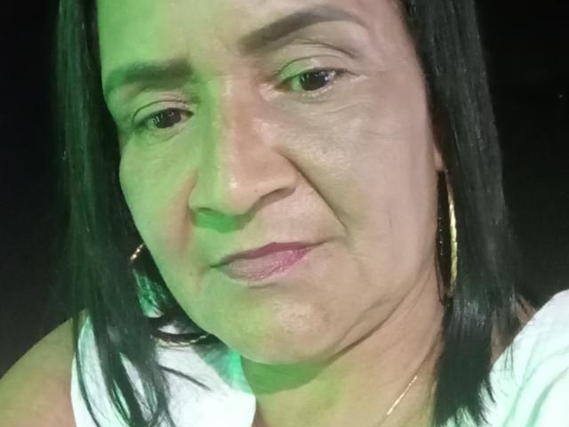 EmillyCam a un visage rond avec un éclairage vert, des yeux marron, des cheveux noirs et lisses, et porte des boucles d'oreilles dorées avec une chemise blanche, affichant un sourire subtil, légèrement laid..